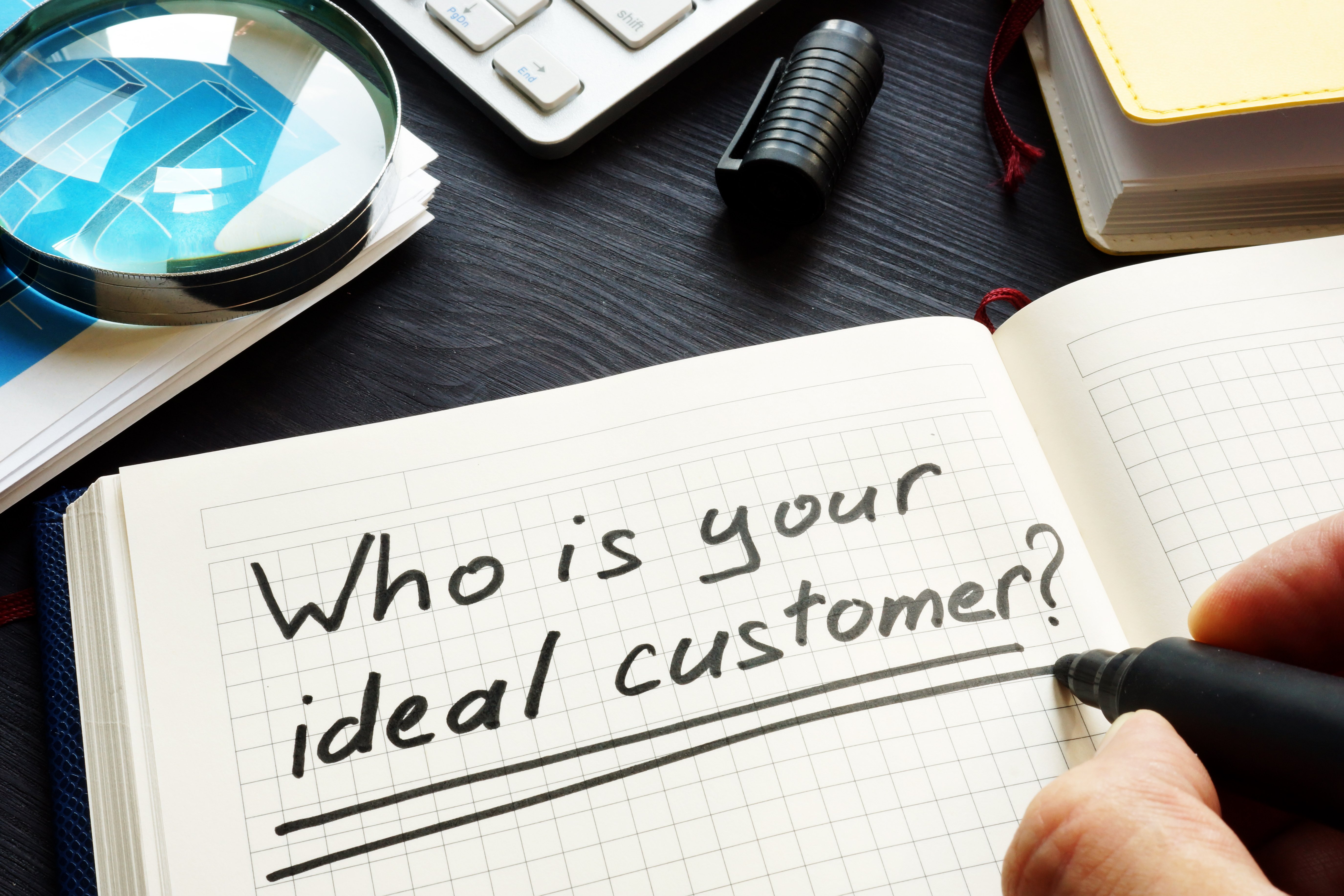 Notebook with handwritten text 'Who is your ideal customer?' next to a magnifying glass, calculator, and office supplies on a desk.