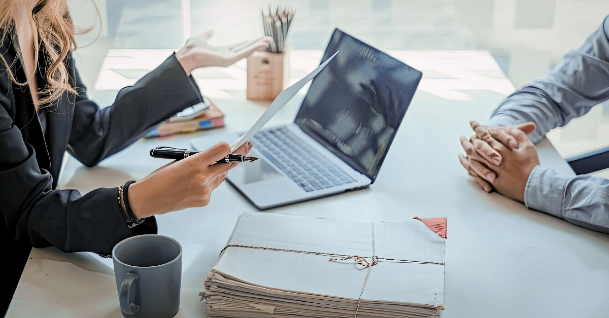 Two people sit at a desk facing each other. On the desk is an open laptop, a stack of files, and a mug. One persons hands are folded while the other holds a pen and appears to be gesturing while speaking. You cannot see their faces in the shot. 