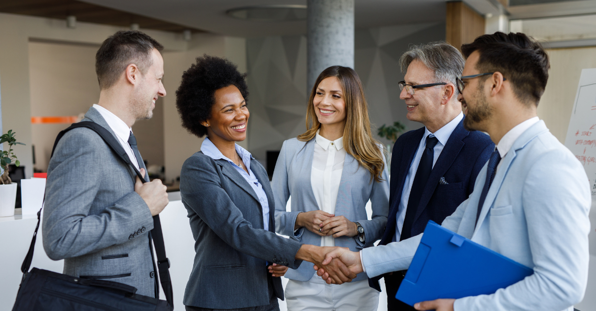 Five people in business attire gather while two shake hands. They appear smiling and in good spirits. 
